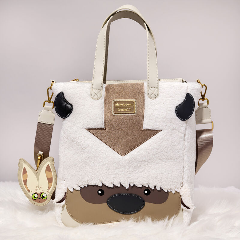 The white sherpa Loungefly Avatar: The Last Airbender Appa Cosplay Plush Tote Bag with Mono Charm sitting on a white furry blanket against a white background. The bag is white sherpa and has appliqué and embroidered details of Appa's face. 